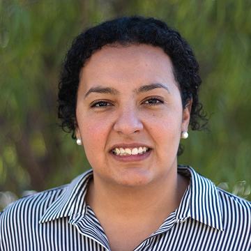Portrait of Mariana Acevedo Nuevo smiling in front of a green leafy background, wearing a striped button-down shirt.